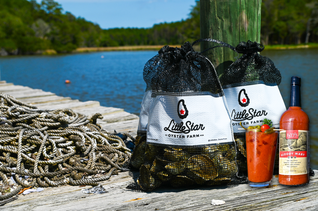 Mesh oyster bags labeled “Little Star Oysters” on a dock by the water with F&TJ'S Bloody Mary Mix