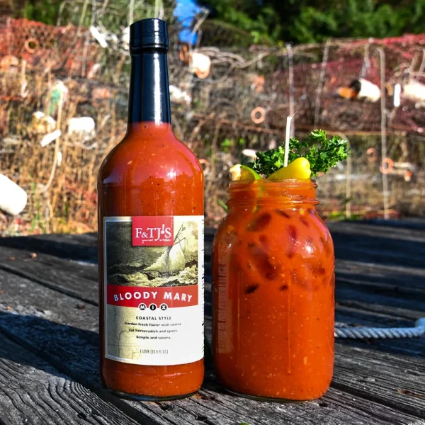 F&TJ'S Carolina Bloody Mary Mix bottle and a garnished mason jar cocktail sitting on a wooden dock with fishing nets in the background.
