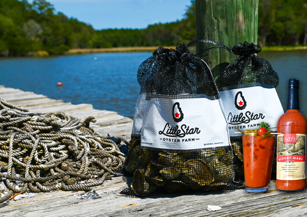 Mesh oyster bags labeled “Little Star Oysters” on a dock by the water with F&TJ'S Bloody Mary Mix