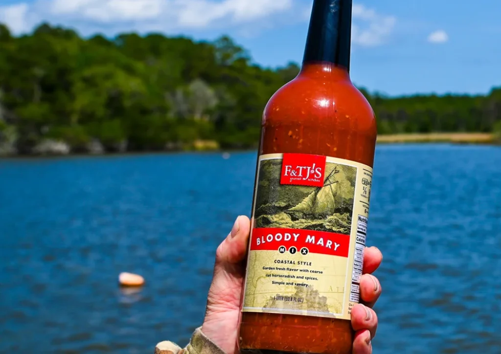 A hand holding a bottle of F&TJ'S Carolina Bloody Mary Mix in front of a scenic river or lake.